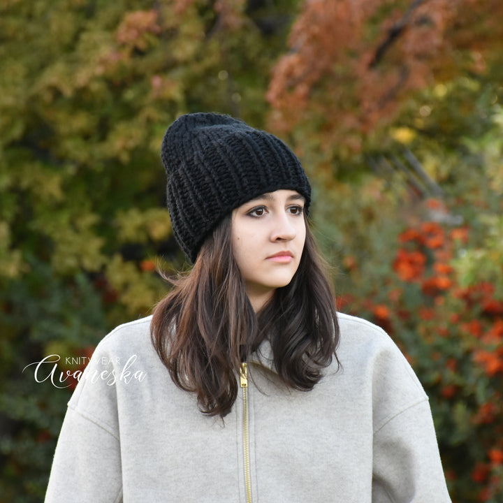 Woman wearing a black knit beanie and gray coat in an outdoor setting with blurred foliage.